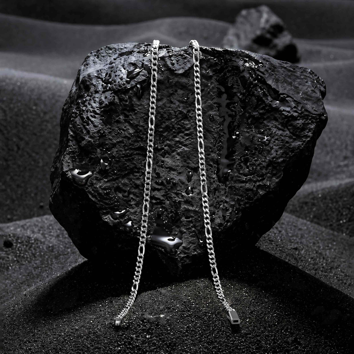 Two silver chain necklaces draped over a dark textured rock, set against a dark, moody background with sand-like textures.