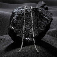 Two silver chain necklaces draped over a dark textured rock, set against a dark, moody background with sand-like textures.