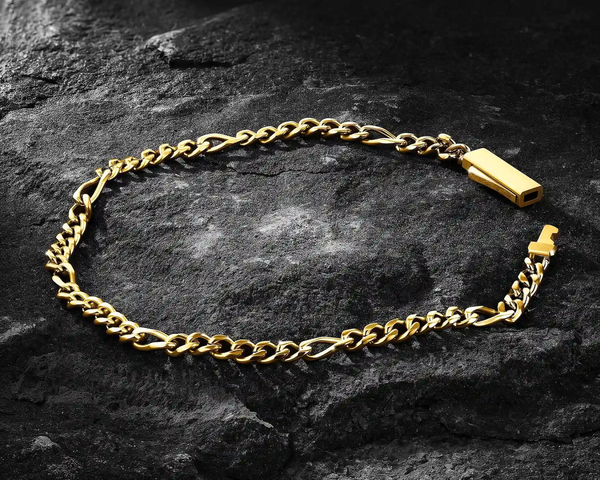 A gold-toned figaro link chain bracelet with a rectangular tag clasp, displayed on a dark textured rock surface.