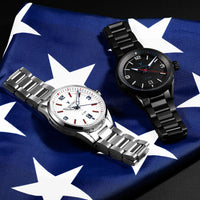Two wristwatches are arranged on a blue-and-white American flag fabric background. The watch on the left is silver-toned with a white dial and red accents, and the watch on the right is black-toned with a black dial and red accents.