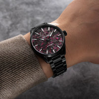 Black wristwatch with red floral pattern on a wrist wearing a brown sweater.