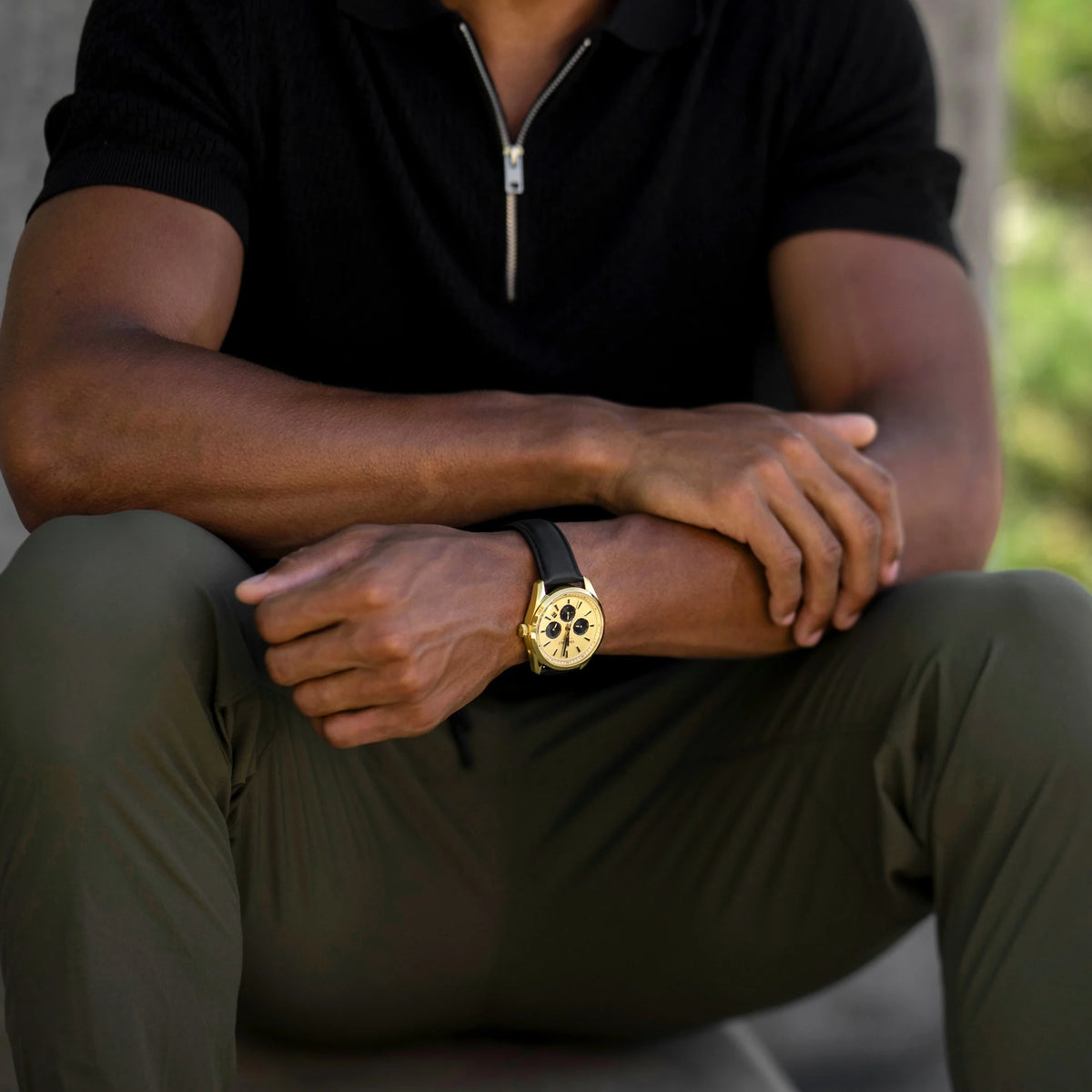 Person wearing a gold watch with a black strap, sitting outdoors.