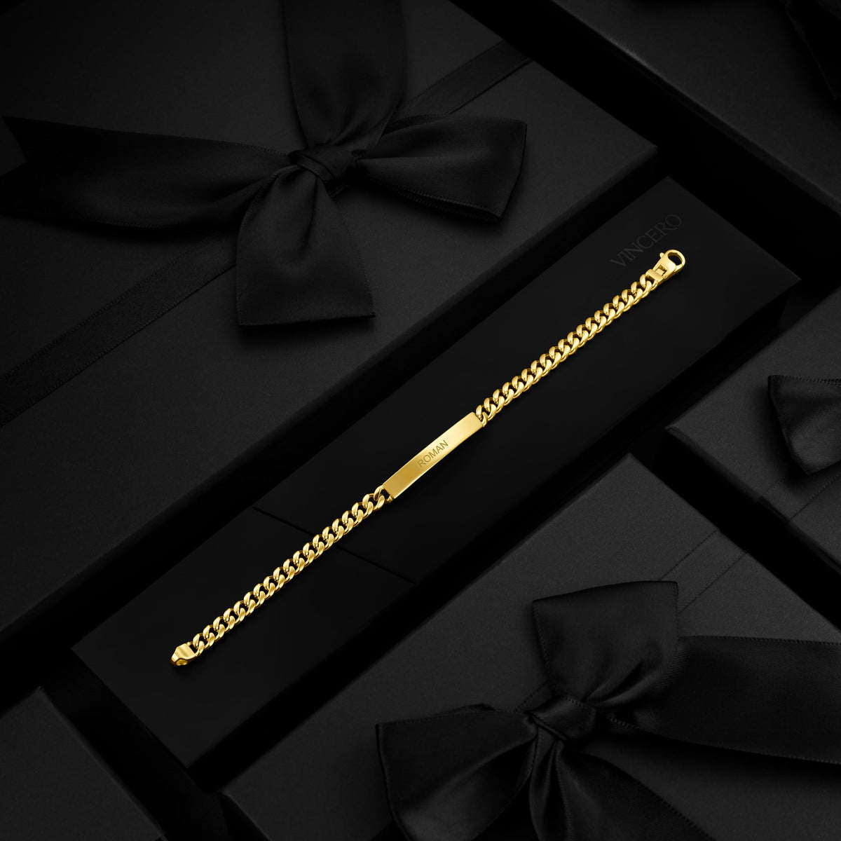 A gold-tone Cuban link chain bracelet with a polished central bar, displayed diagonally on a black presentation box surrounded by black gift boxes and satin bows.