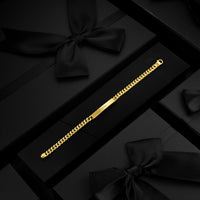 A gold-tone Cuban link chain bracelet with a polished central bar, displayed diagonally on a black presentation box surrounded by black gift boxes and satin bows.