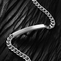 A silver-toned metal chain necklace with a curved bar ID tag, displayed on a dark textured slate background.