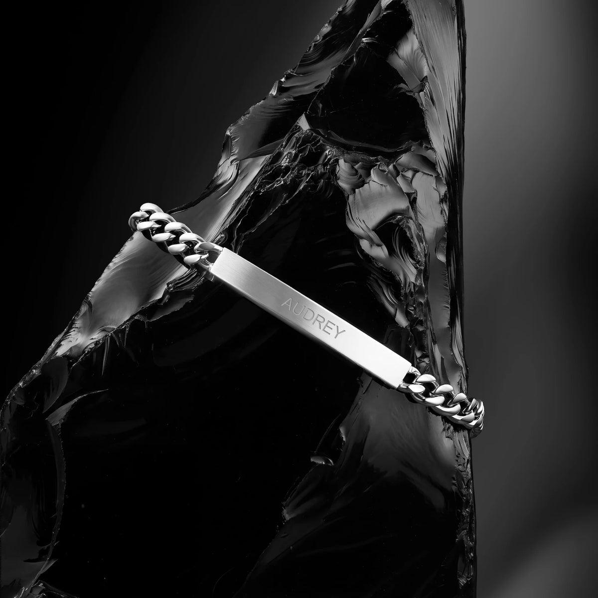 A silver-toned chain bracelet with a polished ID bar engraved with the name 'AUDREY', draped over a glossy black sculptural prop against a dark studio background.