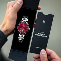Vincero watch with red face and silver band in a black box, held by a person.