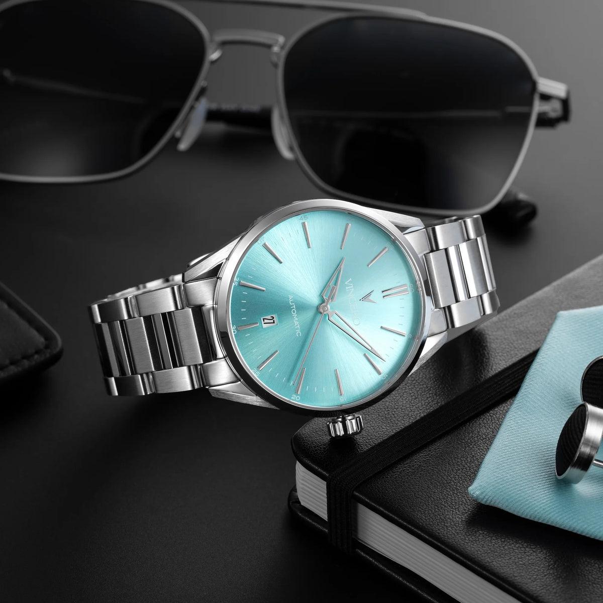Silver watch with turquoise face on a dark surface with sunglasses and a phone case.