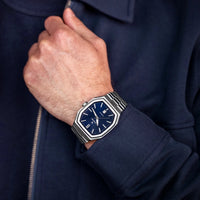 Hand wearing a watch with a dark blue face and silver case, against a matching dark blue background.