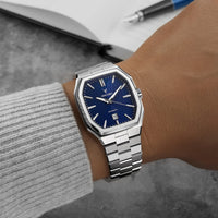 Silver watch with blue dial on a wrist, with a blurred background of a notebook and pen.