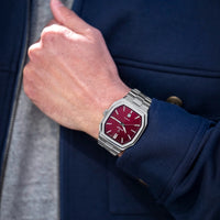 Hand wearing a watch with a red face and silver case, against a dark blue jacket background.