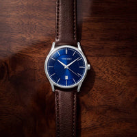 A VINCERO wristwatch with a blue dial and silver case, fitted with a brown leather strap, photographed from above on a dark wood surface.