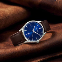 A wristwatch with a blue dial and brown leather strap, resting on a brown leather surface. The watch has a silver-tone case and silver-tone hour markers.