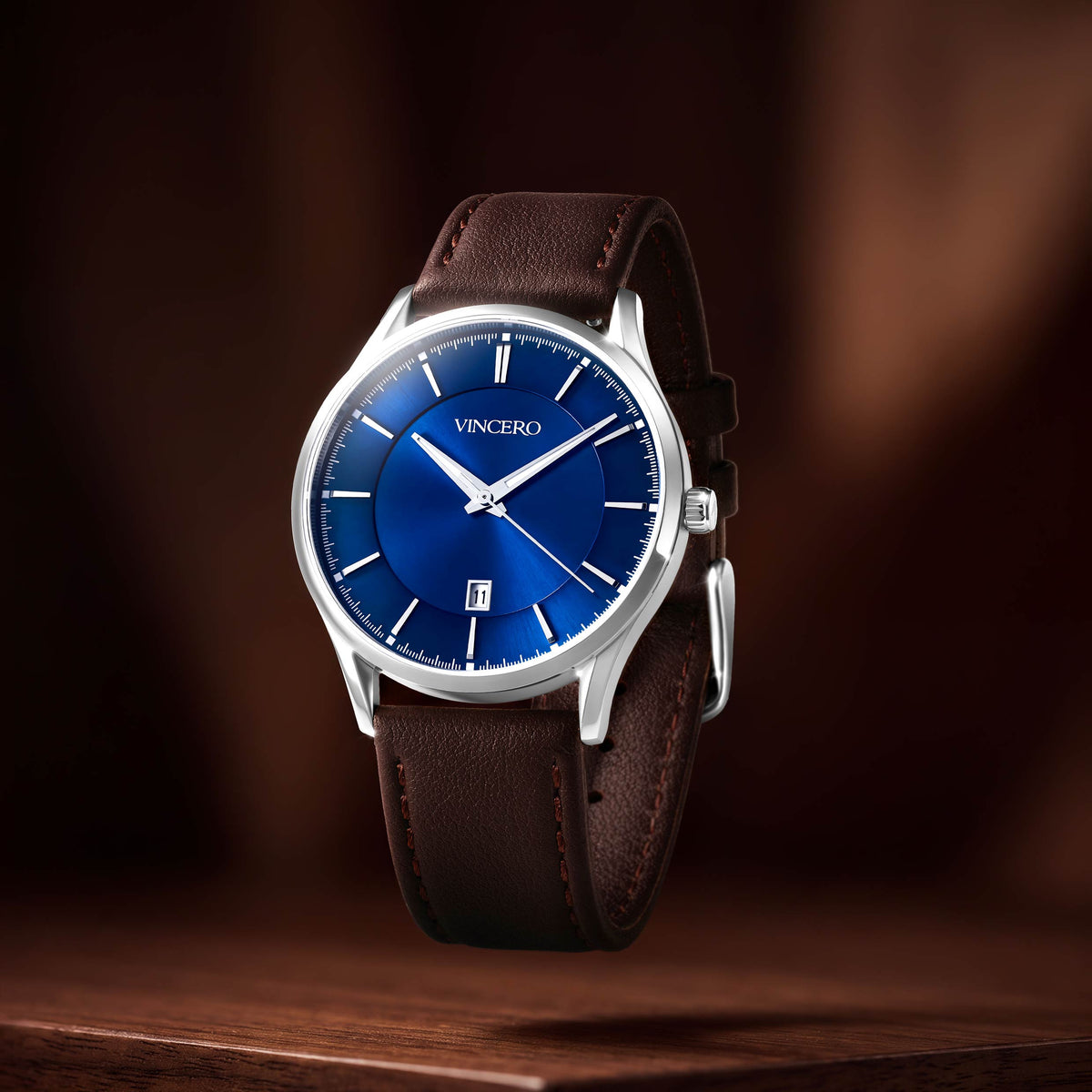 A luxury wristwatch with a vibrant blue dial, silver-tone case, and a dark brown leather strap, shown floating above a wooden surface against a dark brown background.