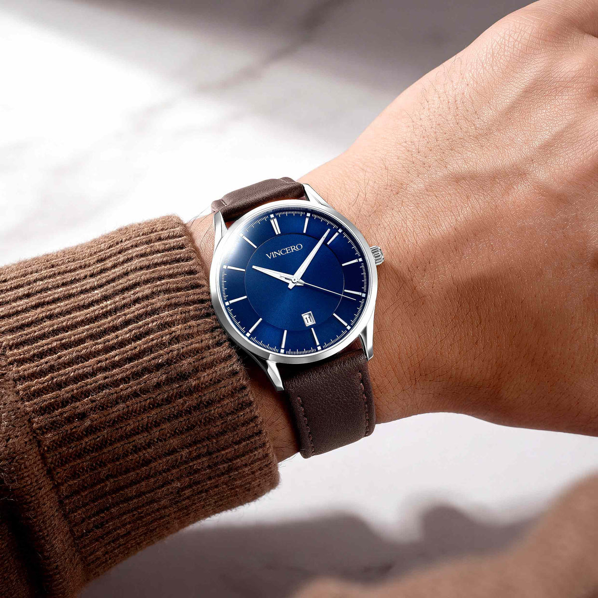 A wristwatch with a deep blue dial and brown leather strap worn on a person's wrist, paired with a brown ribbed sweater sleeve.