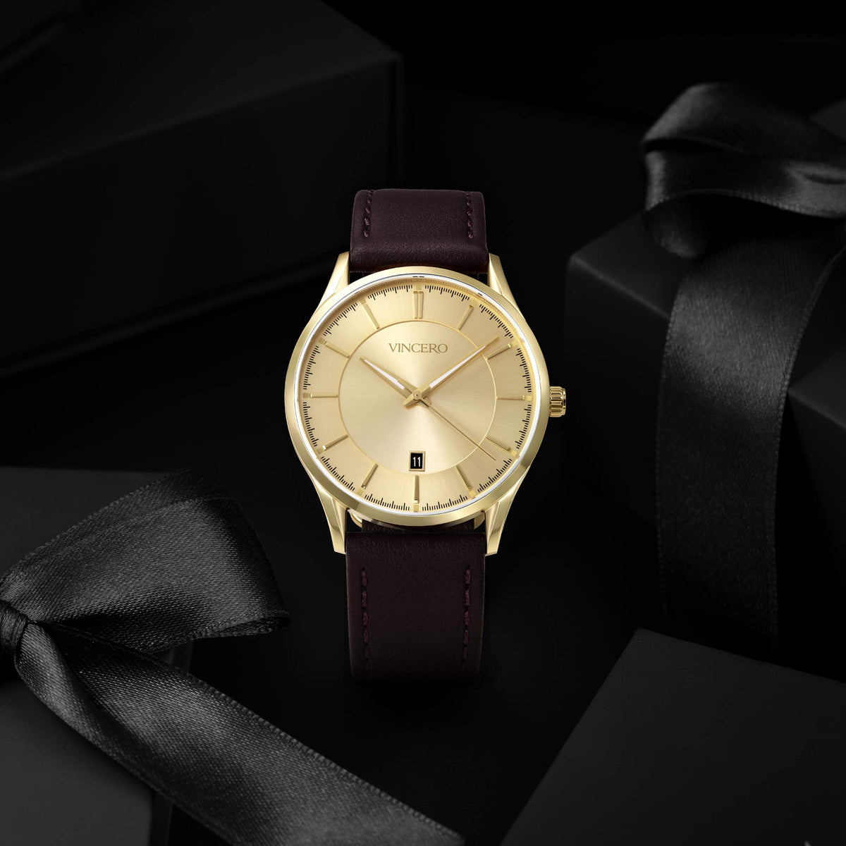A luxury wristwatch by VINCERO with a gold-tone round case, champagne dial, and dark brown leather strap, presented on a black background with gift boxes and satin ribbons.