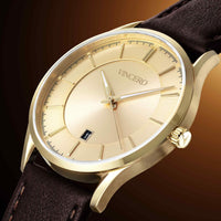 A close-up of a luxury wristwatch with a gold-tone case, matching gold-tone dial, and a dark brown leather strap, shown against a dark brown gradient background.