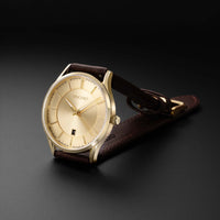 A VINCERO men's watch featuring a gold-tone case, a champagne-gold dial with a date window, and a dark brown leather strap, displayed on a dark gray background.