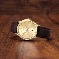 A wristwatch with a gold-toned oval dial, gold-tone case, and dark brown leather strap, resting on a polished wooden surface.