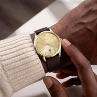 A close-up lifestyle shot of a VINCeRO wristwatch with a gold dial and brown leather strap, worn on a person's wrist and adjusted by another hand.