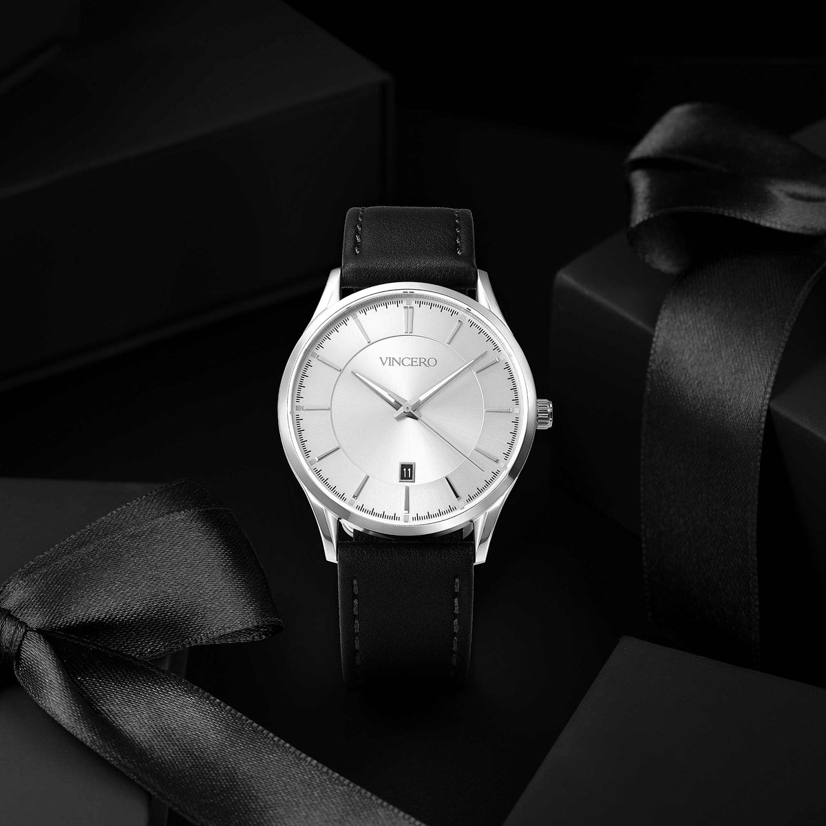 A silver-tone analog wristwatch with a white dial and black leather strap, displayed on a dark surface with black gift boxes and satin ribbons.