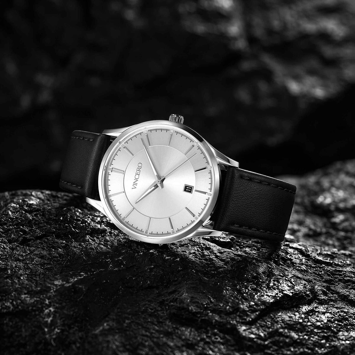 A minimalist wristwatch with a silver-toned round case, white dial, and black leather strap, resting on a dark textured rock surface.