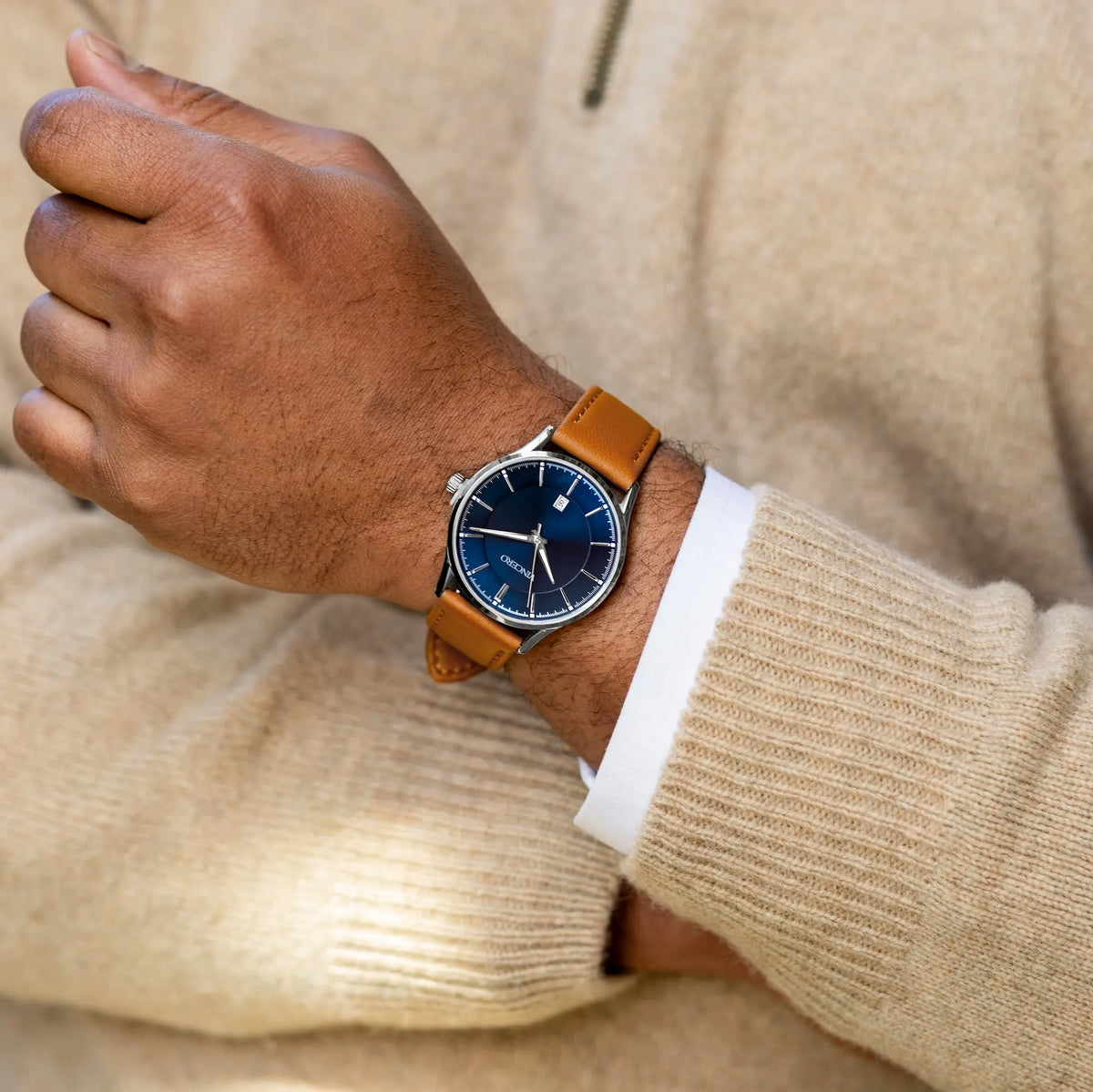 Hand wearing a watch with a brown leather strap and blue dial, against a beige sweater background.