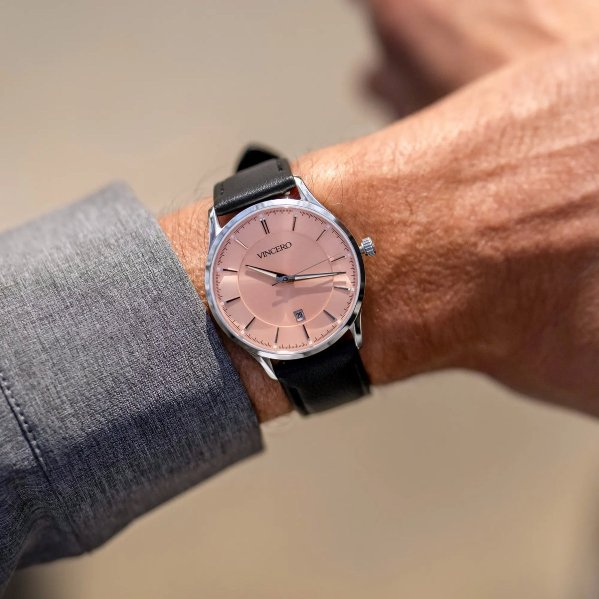 Wrist wearing a watch with a pink face and black strap, on a neutral background.