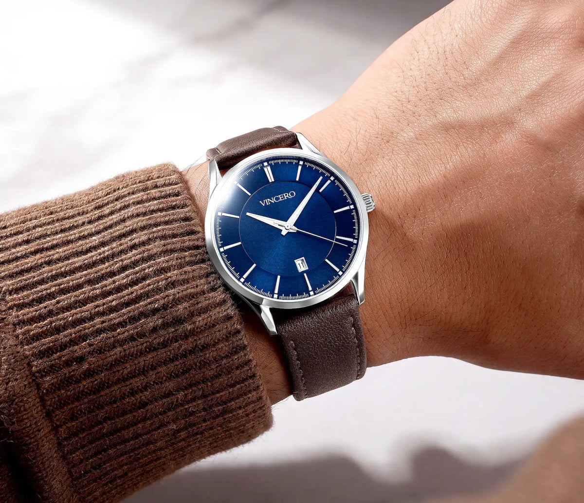 A close-up of a wrist wearing a VINCERO watch with a deep blue dial and a brown leather strap, styled with a brown ribbed knit sweater sleeve.