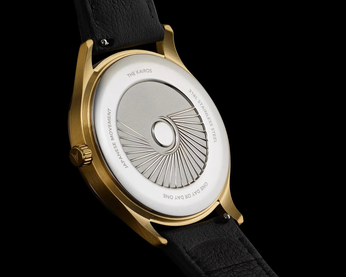 Close-up of the back of a wristwatch with a gold-tone case, black leather strap, and a silver-toned case back featuring engraved sunburst detailing and text including 'THE Kairos'.