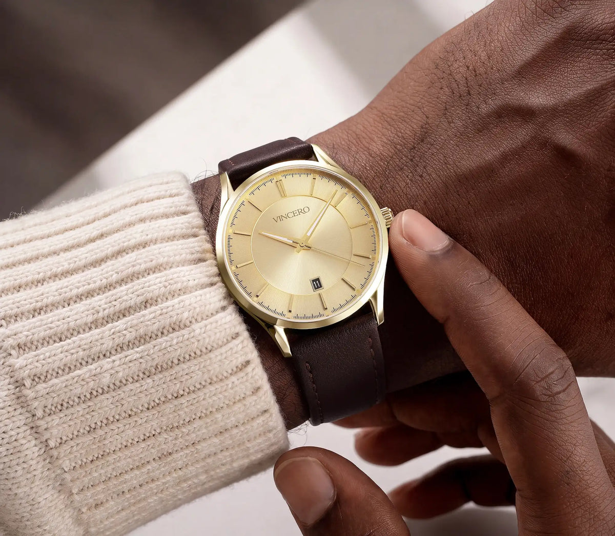 A close-up of a wrist wearing a gold-tone Vincero watch with a dark brown leather strap, styled with a cream ribbed knit sweater sleeve.