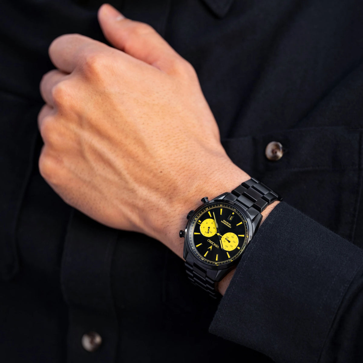 Hand wearing a black watch with yellow accents on a dark background
