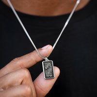 Silver necklace with a rectangular pendant held by a hand against a dark background