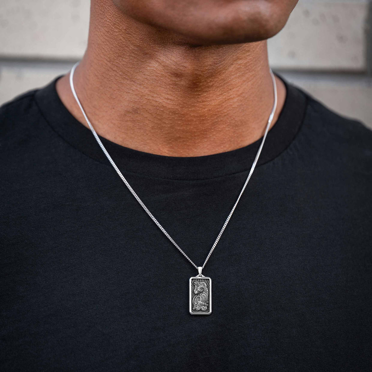 Person wearing a silver necklace with a rectangular pendant on a plain background