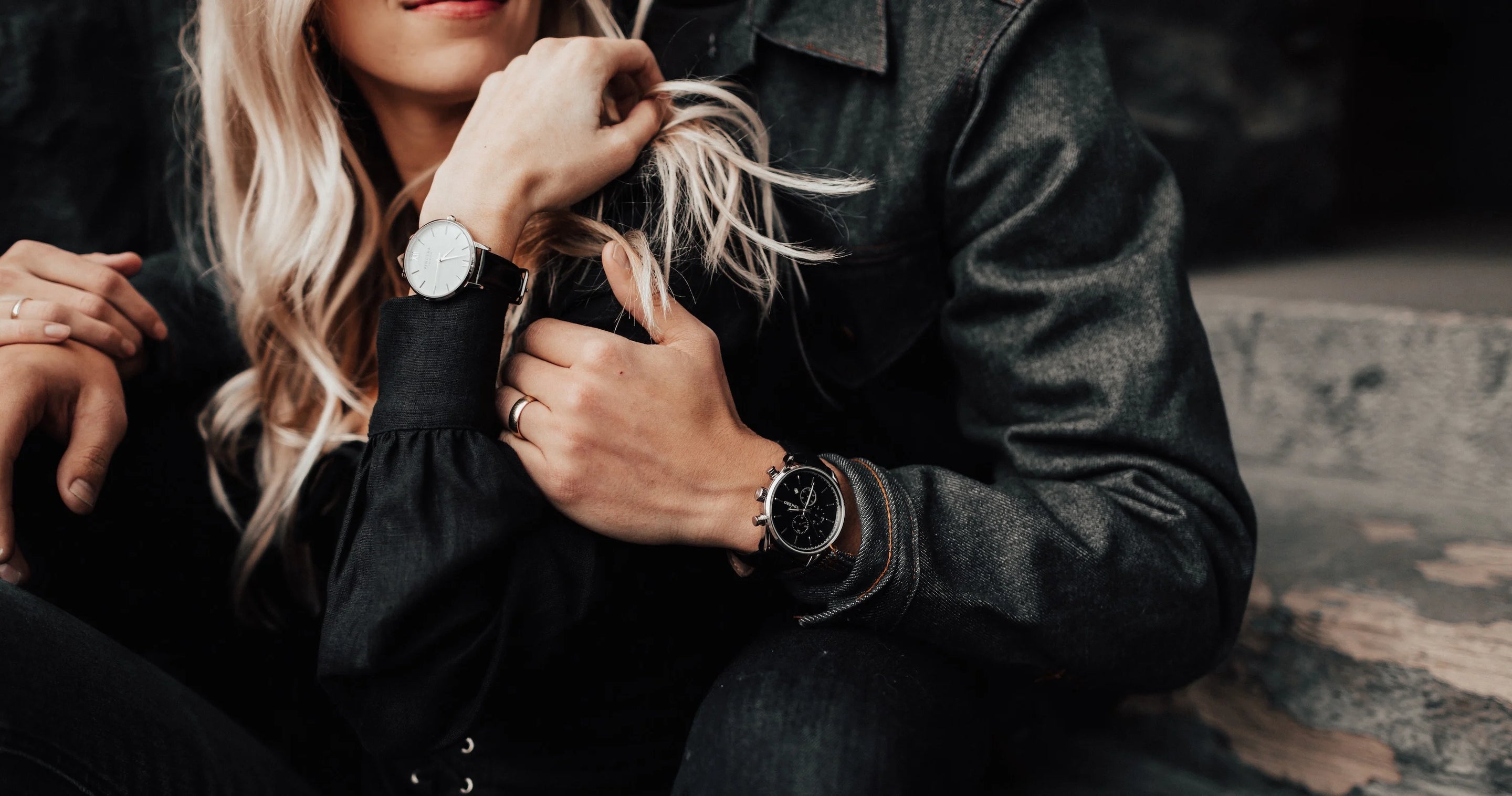 A close-up lifestyle shot of a woman and a man wearing matching black leather jackets and wristwatches. The woman has blonde hair and wears a watch with a white dial and black strap, while the man wears a watch with a black dial and black strap.