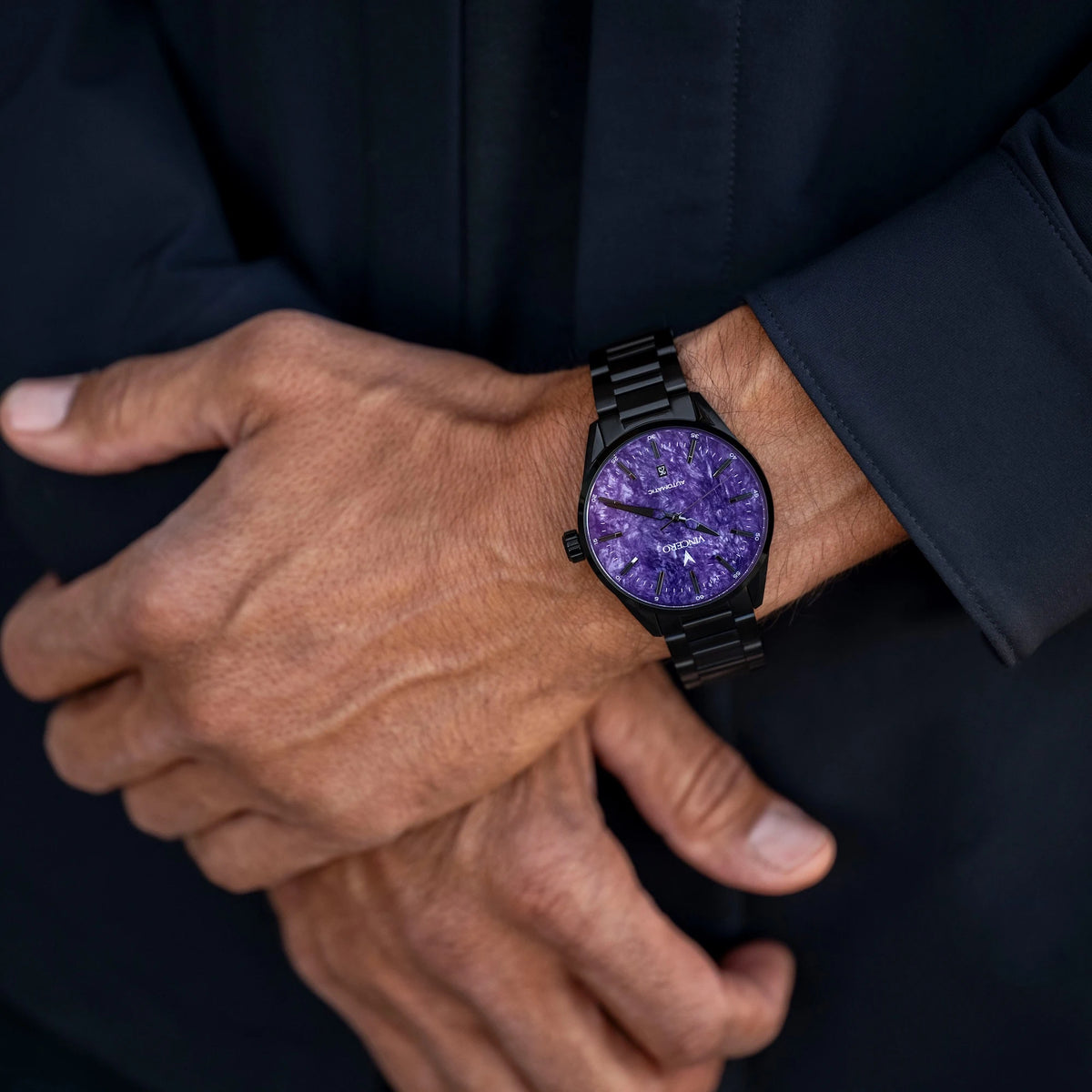 A close-up of a person's hands and wrist wearing a black metal watch with a textured purple dial.