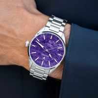 A wristwatch with a silver metal bracelet and case, featuring a textured purple dial, shown worn on a person's wrist against a dark navy suit jacket background.