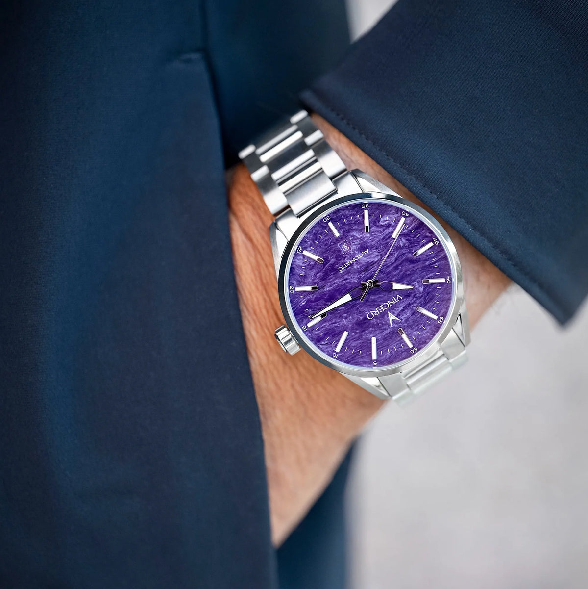 A close-up of a wrist wearing a silver-tone metal bracelet watch with a textured purple dial. The watch is shown on a person's wrist, with a dark navy blazer sleeve visible.