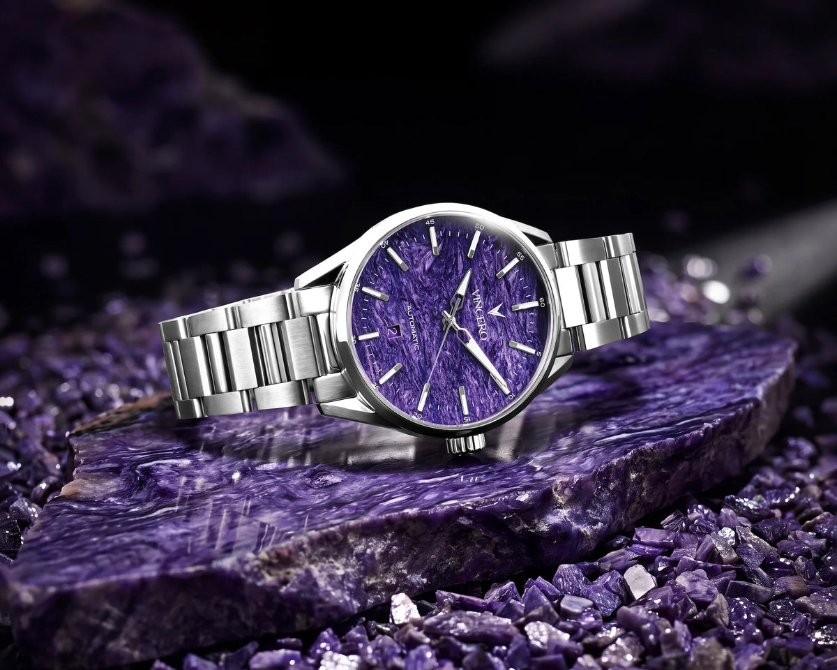 A silver-tone metal wristwatch with a textured purple dial, resting on a polished purple crystal slab with matching purple crystals around it.