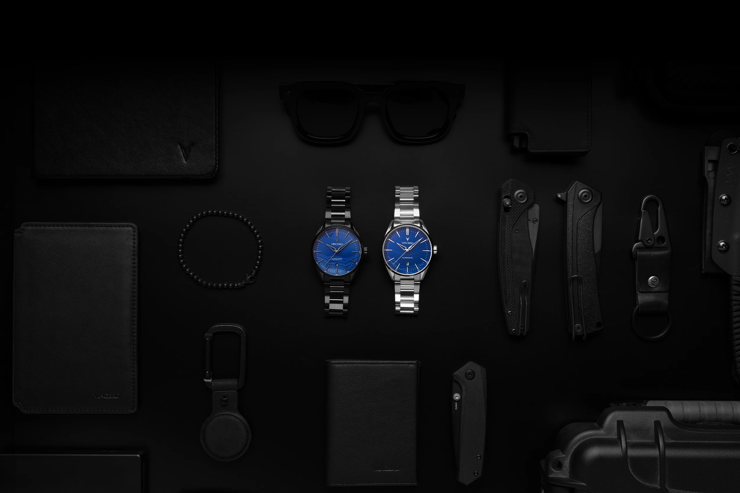 Two blue watches on a dark surface with various items around