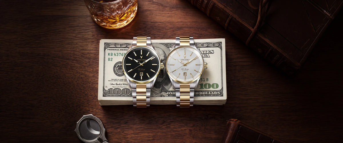 Two wristwatches displayed side by side on top of a stack of $100 bills, set on a dark wooden table with a glass of whiskey, a leather portfolio, and a cigar case nearby.