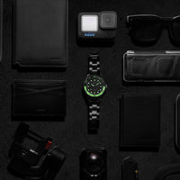 Collection of black and green items including a watch, camera, and sunglasses on a dark surface.