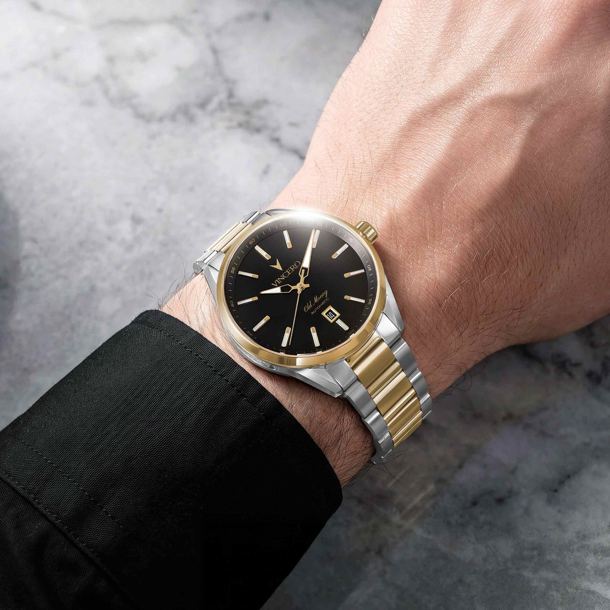 A two-tone wristwatch with a black dial and gold-tone markers, shown worn on a person's wrist. The watch features a gold-tone case, a silver-tone and gold-tone link bracelet, and a date window at 3 o'clock.