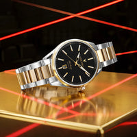 A luxury wristwatch with a black dial, gold-tone markers and hands, and a two-tone silver and gold metal bracelet, displayed on a reflective gold surface with red laser lines in the background.