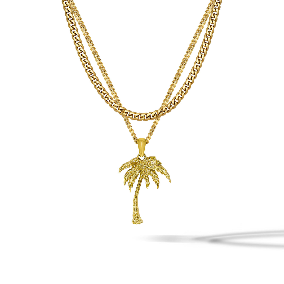 A gold-toned double-strand necklace featuring a curb chain and a thinner chain with a detailed palm tree pendant, displayed on a clean white background with a soft shadow.