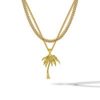 A gold-toned double-strand necklace featuring a curb chain and a thinner chain with a detailed palm tree pendant, displayed on a clean white background with a soft shadow.