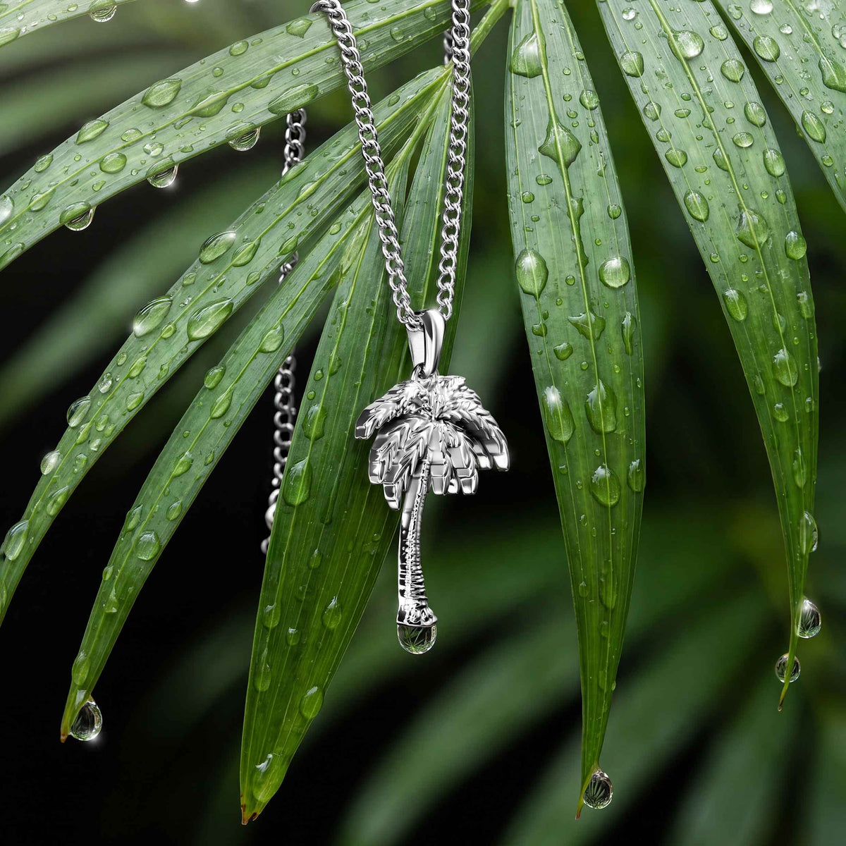 A silver palm tree pendant necklace hanging from a silver chain, set against green palm leaves covered in water droplets.
