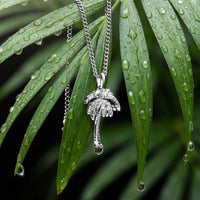 A silver palm tree pendant necklace hanging from a silver chain, set against green palm leaves covered in water droplets.