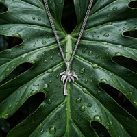 A silver-toned necklace with a detailed palm tree pendant, displayed on a large green monstera leaf covered in water droplets.