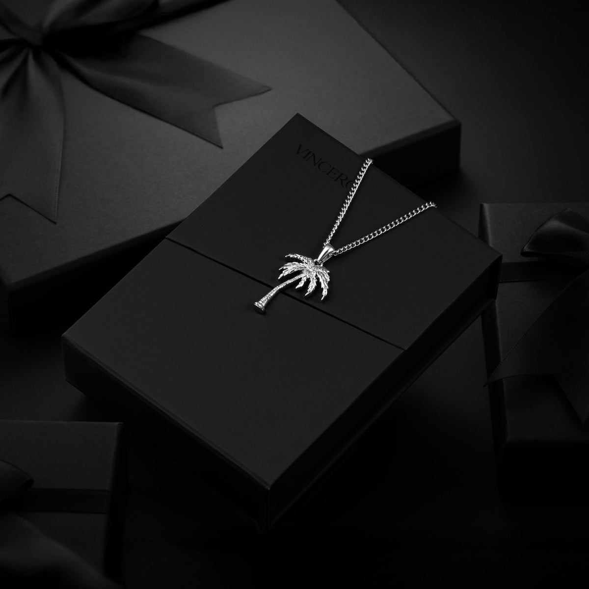 A silver-toned necklace with a palm tree pendant displayed on a black gift box, surrounded by dark gift packaging and ribbons.